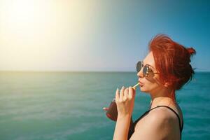 ragazza su estate vacanza. dai capelli rossi donna nel occhiali da sole bevande limonata a partire dal bottiglia attraverso cocktail tubo. blu mare, luce del sole e chiaro cielo su sfondo. copia spazio per viaggio agenzia pubblicità. foto