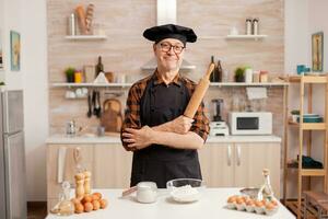 anziano età uomo indossare capocuoco bonete sorridente nel casa cucina. pensionato panettiere nel cucina uniforme preparazione Pasticcino ingredienti su di legno tavolo pronto per cucinare fatti in casa gustoso pane, torte e pasta foto