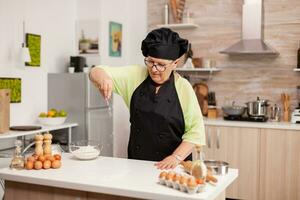 panettiere diffusione Farina su di legno tavolo a casa nel moderno cucina. contento anziano capocuoco con uniforme aspersione, setacciare vagliatura crudo ingredienti di mano cottura al forno fatti in casa Pizza foto