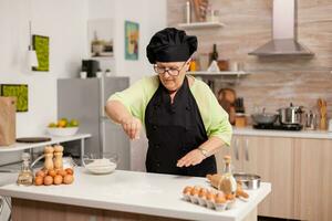 anziano capocuoco con uniforme aspersione Farina nel casa cucina indossare grembiule e ossatura. contento anziano capocuoco con uniforme aspersione, setacciare vagliatura crudo ingredienti di mano cottura al forno fatti in casa Pizza. foto