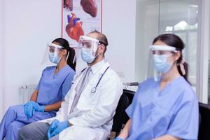 stanco medici con viso maschera e visiera contro coronavirus infezione seduta nel ospedale ricezione la zona guardare a telecamera. professionale medico personale Lavorando nel nuovo normale ospedale ufficio foto