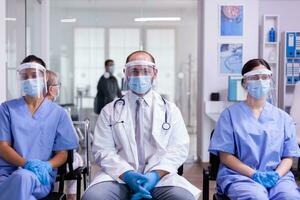 gruppo di donne infermieri e uomo medico nel laboratorio cappotti e scrub guardare a telecamera indossare viso maschera e visiera contro coronavirus seduta nel ospedale ricezione la zona. pazienti rispetto sociale distanza foto