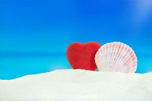 rosso cuore con conchiglia su sabbia di spiaggia dietro a mare. San Valentino giorno, nozze, luna di miele, vacanza nel caldo Paesi. copia spazio foto