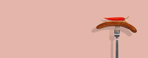 banner con salsiccia alla griglia singola con senape su di esso e peperoncino rosso piccante messo su una forchetta moderna in metallo su uno sfondo di colore rosa solido con spazio di copia per il testo. concetto di cibo di strada e cucina. foto