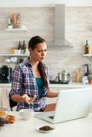 donna guardare confuso a il computer portatile schermo mentre Lavorando a partire dal casa nel cucina. Lavorando a partire dal casa utilizzando dispositivo con Internet tecnologia, navigando, ricerca su aggeggio nel il mattina. foto