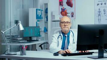 anziano medico nel moden privato clinica Lavorando a pc poi sorridente a il telecamera. assistenza sanitaria specialista medicina professionista nel uniforme Lavorando nel ospedale con grande personale foto