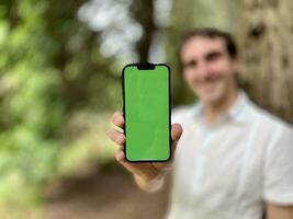 sfocato messa a fuoco solo Telefono nel messa a fuoco molto bello uomo francese Italia non Tenere un' Telefono con un' verde rubinetto in piedi nel il boschi nel un' bianca camicia e sorridente mostrando un' dito su il schermo può uso per foto