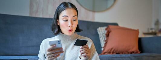 ritratto di sorridente asiatico donna pagare con credito carta su sua smartphone app, organizzare diretto addebito o shopping in linea a partire dal mobile Telefono foto
