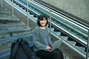 ritratto di bellissimo giovane coreano donna, alunno si siede su le scale nel pubblico posto, ascolta musica nel cuffia, lavori su progetto su computer portatile, lavori remoto foto