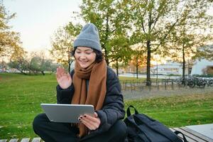 ritratto di giovane asiatico ragazza si siede nel caldo Abiti nel parco, onde mano a tavoletta, video chat all'aperto, dice Ciao mentre su chiamata foto