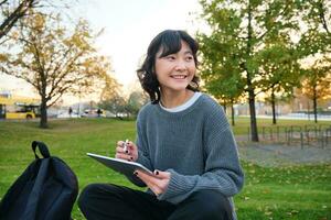 giovane coreano donna si siede nel parco, disegna su digitale tavoletta, scrive nel aggeggio con penna attrezzo, sembra in giro e fa Appunti, crea opera d'arte o graffi foto