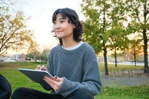 ritratto di giovane sorridente coreano ragazza, grafico progettista, artista disegno su digitale tavoletta con un' penna attrezzo, seduta nel parco su fresco aria e graffiare, assunzione Appunti foto