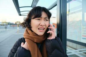 giovane donna sembra frustrato e deluso, parla su mobile Telefono, sta su autobus fermare, riceve cattivo notizia foto