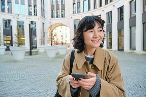 elegante coreano ragazza nel cuffia, ascolta musica e usi mobile Telefono, sta nel città centro, aspetta per qualcuno su strada e scrive testo Messaggio su sua smartphone foto