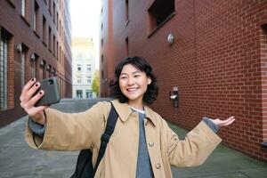 contento coreano ragazza, turista prende fotografie su strada, Spettacoli smth mentre record video di se stessa su smartphone, in posa vicino edificio e sorridente