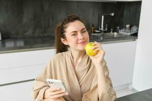 vicino su ritratto di contento giovane donna nel accappatoio, seduta nel il cucina e utilizzando mobile Telefono, Tenere un mela, ordine frutta e verdure in linea, utilizzando smartphone App per drogheria consegna foto