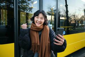 entusiasta asiatico donna, in piedi su autobus fermare con smartphone, guardare a Telefono schermo con stupito, trionfante viso, vincente, sentire grande notizia su video Chiacchierare foto