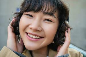 ritratto di contento donna, coreano ragazza nel cuffia, ascoltando musica nel cuffia, godendo suono di auricolari, ridendo e sorridente foto