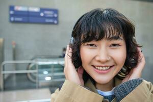 vicino su ritratto di elegante coreano ragazza in piedi su strada, ascoltando musica nel cuffie e sorridente, in posa nel città centro foto