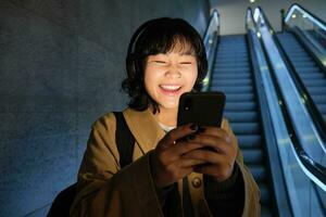 ridendo carino coreano ragazza su scala mobile, andando giù per tubo, metropolitana o la metropolitana, utilizzando mobile Telefono, Guardando smth divertente su smartphone App foto