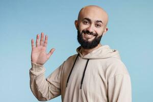 allegro arabo uomo agitando Ciao con mano e guardare a telecamera. contento giovane Calvo barbuto persona con amichevole facciale espressione indossare casuale Abiti saluto con braccio studio ritratto foto