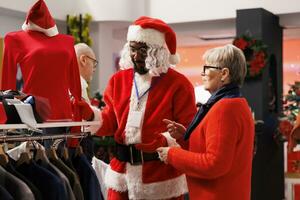 uomo nel Santa costume mostrando elementi su cremagliere per cliente, anziano donna chiede dipendente di dimensioni a disposizione nel negozio. gioviale persona nel rosso completo da uomo porzione cliente per trova natale regalo. foto