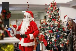 gioioso lavoratore vestito come Santa Claus nel Natale decorato shopping centro, attrarre i clienti nel capi di abbigliamento negozio. dipendente tintinnio campane e saluto clienti nel festivo moda negozio. foto