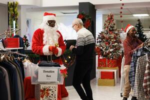 uomo nel Santa completo da uomo mettendo donazione scatola nel negozio, incoraggiando i clienti per donare Abiti per beneficenza su Natale vacanza. Al dettaglio lavoratore nel costume e clienti la donazione merce per bambini. foto