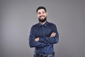 giovane bell'uomo appoggiato al muro grigio con le braccia incrociate. un giovane serio con la barba guarda la telecamera foto