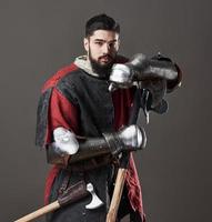 cavaliere medievale su sfondo grigio. ritratto di brutale guerriero faccia sporca con armatura di cotta di maglia vestiti rossi e neri e ascia da battaglia