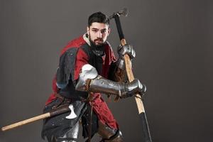 cavaliere medievale su sfondo grigio. ritratto di brutale guerriero faccia sporca con armatura di cotta di maglia vestiti rossi e neri e ascia da battaglia foto