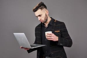 Occupato uomo barbuto in abiti da lavoro che parla tramite smartphone e utilizza il computer portatile mentre si tiene una tazza di caffè in mano su sfondo grigio foto