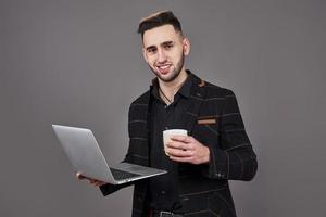 Occupato uomo barbuto in abiti da lavoro che parla tramite smartphone e utilizza il computer portatile mentre si tiene una tazza di caffè in mano su sfondo grigio foto