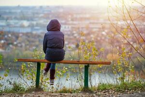 giovane donna seduta su panchina nel autunno città parco e guardare a paesaggio urbano, indietro Visualizza foto