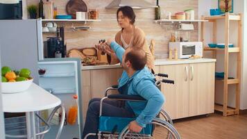 Disabilitato uomo nel sedia a rotelle porzione moglie nel cucina. apertura frigorifero. tipo con paralisi handicap invalidità portatori di handicap le difficoltà ottenere Aiuto per mobilità a partire dal amore e relazione foto