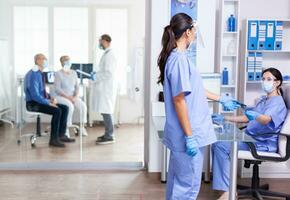 medico infermiera Tenere raggi X nel ospedale corridoio durante coronavirus scoppio indossare viso maschera . assistente Lavorando su ricezione computer. medico discutere con anziano coppia foto