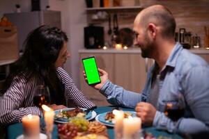 coppia Guardando Telefono con verde schermo. contento uomo e donna guardare a verde schermo modello croma chiave isolato smartphone Schermo utilizzando tecnologia Internet seduta a il tavolo nel cucina. foto