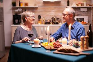 avendo un' conversazione durante frastuono romantico cena. contento allegro anziano anziano coppia cenare insieme nel il accogliente cucina, godendo il pasto, festeggiare loro anniversario. foto