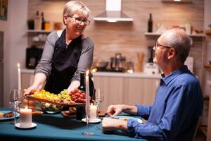 anziano donna servendo marito con uva e noccioline come aperitivo per romantico cena. anziano vecchio coppia parlando, seduta a il tavolo nel cucina, godendo il pasto, festeggiare loro anniversario . foto