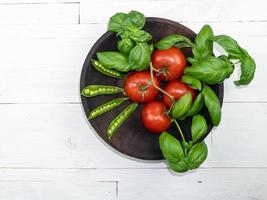 pomodori, basilico e baccelli di piselli verdi in un piatto scuro su un tavolo di legno foto