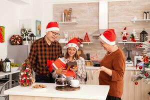 contento nonni sorprendente nipotina con natale presente regalo con nastro su esso durante periodo natalizio nel x-mas decorato cucina. famiglia godendo inverno stagione festeggiare Natale vacanza foto