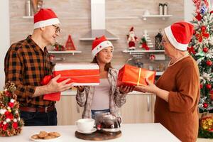 contento famiglia portando natale involucro regalo con nastro su esso festeggiare Natale vacanza nel x-mas decorato cucina godendo inverno stagione. nonni sorprendente bambino con Santa Claus presente foto