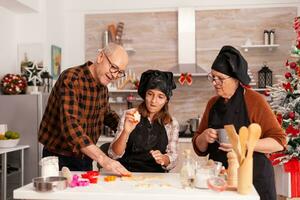 contento famiglia cucinando tradizionale Pan di zenzero dolce fabbricazione Impasto utilizzando cottura al forno albero forma nel natale decorato cucina. nipote festeggiare Natale vacanza insieme preparazione fatti in casa biscotti foto