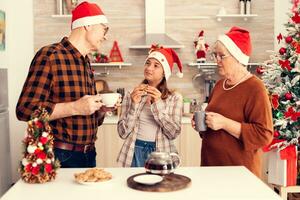 nipote bonding con nonna festeggiare Natale godendo gustoso biscotti durante prima colazione. contento bambino indossare Santa cappello mangiare delizioso biscotti a partire dal nonno mentre festeggiare inverno vacanze con loro. foto