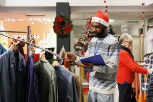 Al dettaglio lavoratore fare elementi inventario con appunti nel mano, indossare Santa cappello nel capi di abbigliamento memorizzare decorato per Natale. shopping assistente conteggio tutti merce e Abiti a partire dal grucce. foto