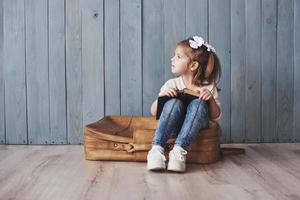 pronto per il grande viaggio. bambina felice che legge un libro interessante che trasporta una grande valigetta e sorride. concetto di viaggio, libertà e immaginazione foto