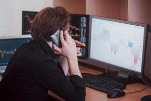 vista da sopra e da un broker di borsa che fa trading online mentre accetta ordini per telefono. più schermi di computer pieni di grafici e analisi dei dati in background foto