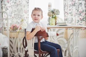 piccolo ragazzo in jeans pantaloni blu è seduto sulla sedia di legno nella stanza. fa i compiti. di nuovo a scuola foto