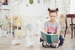 carina bambina che legge un libro in camera da letto. bambino con la corona seduto sul letto vicino alla finestra foto