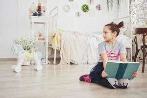 carina bambina che legge un libro in camera da letto. bambino con la corona seduto sul letto vicino alla finestra foto